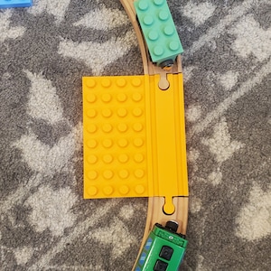 May include: A wooden train track set with a green toy train and a yellow and orange bridge. The train track is on a gray carpet.