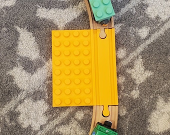 May include: A wooden train track set with a green toy train and a yellow and orange bridge. The train track is on a gray carpet.
