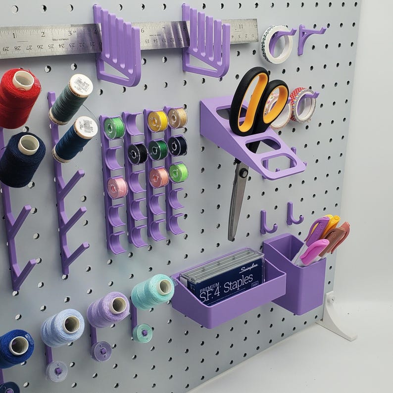 Puede incluir: Un tablero de clavijas morado con varias herramientas y suministros para manualidades y costura. Hay carretes de hilo, una regla, tijeras, un portagrapas y un peque&ntilde;o contenedor para bol&iacute;grafos y l&aacute;pices. El tablero de clavijas est&aacute; hecho de pl&aacute;stico y tiene un patr&oacute;n de cuadr&iacute;cula.