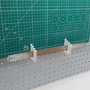 Op de afbeelding: Witte kunststof haken voor een prikbord, gemonteerd op een grijze prikbordwand. Een liniaal hangt aan de haken.