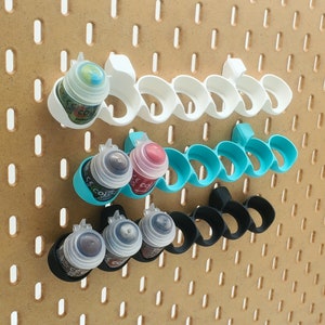 Puede incluir: Un conjunto de seis soportes para botes de pintura, hechos de plástico blanco, azul y negro. Los soportes están diseñados para sujetar botes de pintura pequeños y están dispuestos en fila en una tabla de clavijas.