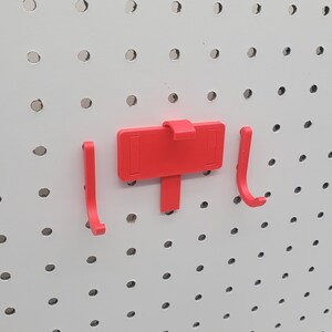 May include: Three red plastic pegboard hooks. Two hooks are shaped like a 'J' and one hook is a flat rectangular shape with a small hook on the top.