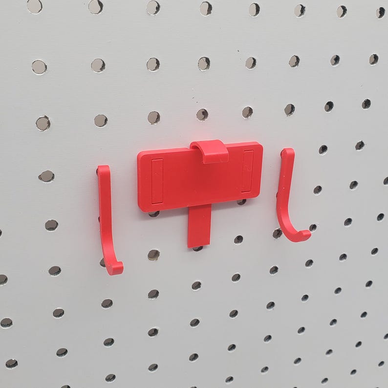 May include: Three red plastic pegboard hooks. Two hooks are shaped like a 'J' and one hook is a flat rectangular shape with a small hook on the top.