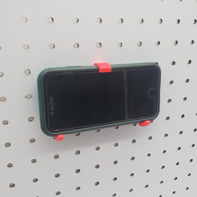 May include: A black smartphone with a green case is mounted on a white pegboard using a red plastic phone holder.