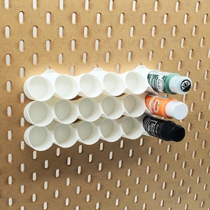 May include: A white plastic paint bottle organiser mounted on a pegboard. The organiser holds three rows of paint bottles. Three paint bottles are in the organiser, with green, orange, and black paint. The pegboard is a light brown colour.
