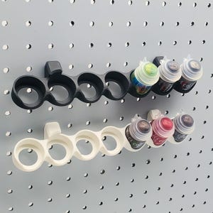 Puede incluir: Dos soportes para botes de pintura, uno negro y otro blanco, están montados en una pared de tablero perforado. Cada soporte tiene seis ranuras para botes de pintura. El soporte negro tiene tres botes de pintura, y el soporte blanco tiene tres botes de pintura.