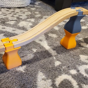 Könnte beinhalten: Ein Holzbahn-Set mit drei orangefarbenen und grauen Stützen. Die Bahn ist gebogen und die Stützen sind vasenförmig. Die Bahn befindet sich auf einem grauen Teppich.