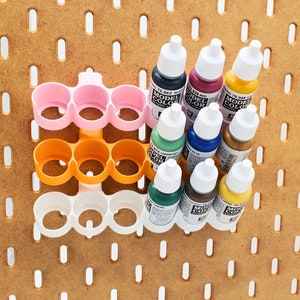 May include: A white, orange, and pink plastic paint bottle holder with three rows of three bottle holders. The holder is filled with eight bottles of acrylic paint. The bottles are labeled "Model Color" and have different colors, including black, red, yellow, green, blue, and brown.