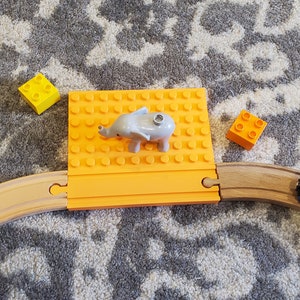 May include: A toy train set with a wooden track, orange and yellow blocks, and a gray elephant figurine on an orange platform. The train cars are red and yellow.