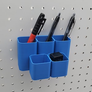 May include: A set of six blue plastic wall mounted organizers with three pens and a small black box. The organizers are mounted on a white pegboard.