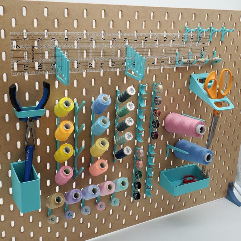 Puede incluir: Un tablero de clavijas con varias herramientas y suministros para coser y manualidades. El tablero de clavijas es marr&oacute;n con una cuadr&iacute;cula de agujeros. Hay varios ganchos y soportes de color turquesa, as&iacute; como carretes de hilo de varios colores. Hay una regla, unas tijeras y un peque&ntilde;o contenedor de color turquesa.