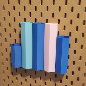 Puede incluir: Cinco contenedores de almacenamiento hexagonales en colores azul, azul claro, rosa y azul oscuro están montados en una pared de tablero de clavijas.