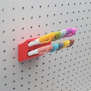 May include: A red plastic wall-mounted organizer with two slots for holding markers. The organizer is mounted on a white pegboard.  The organizer is holding eight markers in various colors, including yellow, orange, pink, blue, green, and purple.