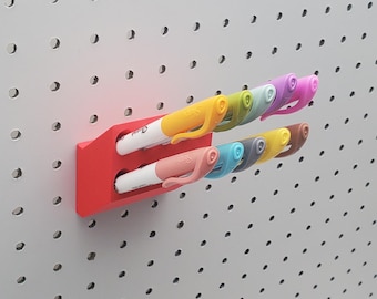 May include: A red plastic wall-mounted organiser with two slots for holding markers. The organiser is mounted on a white pegboard.  The organiser is holding eight markers in various colours, including yellow, orange, pink, blue, green, and purple.