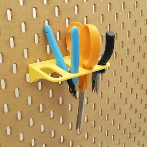 May include: A yellow plastic wall-mounted tool holder with three slots holds a pair of blue and yellow scissors, a pair of blue pliers, and a pair of silver pliers.