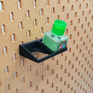 Op de afbeelding: Een zwarte plastic pegboardhaak met een klein flesje groene verfverdunner erin. De fles heeft een wit etiket met de tekst "Tamiya Extra Thin Cement" erop gedrukt.