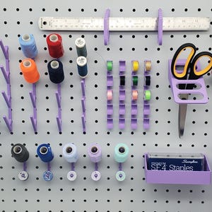Puede incluir: Un tablero de clavijas con varias herramientas y suministros para manualidades y costura. Hay carretes de hilo de diferentes colores, una regla, tijeras, cinta adhesiva y un contenedor para grapas. El tablero de clavijas es morado y tiene un fondo blanco.