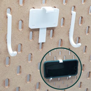 May include: White plastic hooks and a phone holder mounted on a pegboard wall. The phone holder is holding a black smartphone.