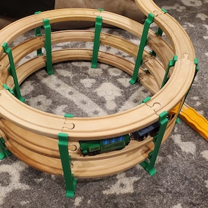 Puede incluir: Juego de vías de tren de juguete de madera con un diseño en espiral. La vía es de madera marrón claro y está sostenida por postes de plástico verde. Dos trenes de juguete están en la vía y una rampa amarilla está conectada.