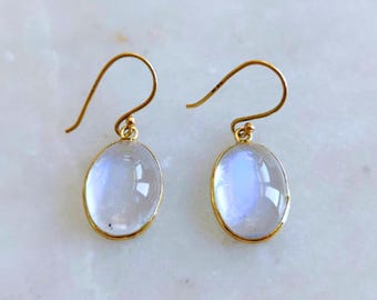 May include: A pair of gold-colored earrings with oval, iridescent, moonstone-like gems. The gems are set in a gold bezel and have a subtle blue and white color. The earrings have a simple hook design.