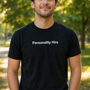 Puede incluir: Camiseta negra de cuello redondo con el texto blanco "Personality Hire". La persona que lleva la camiseta está de pie afuera con un fondo verde borroso.
