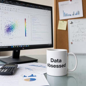 May include: A white ceramic mug with the words "Data obsessed" in black lettering. The mug sits on a desk with a computer monitor displaying data visualizations, a calculator, and printed charts. A corkboard and whiteboard are in the background.