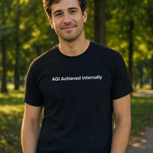 Puede incluir: Una camiseta negra con el texto blanco "AGI Achieved Internally". La persona que lleva la camiseta está de pie en un parque con árboles al fondo. La persona está sonriendo.