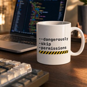 May include: A white ceramic mug featuring the black text --dangerously -skip -permissions above a yellow and black hazard warning stripe. The mug has a standard handle and a cylindrical body, positioned in front of a laptop computer.