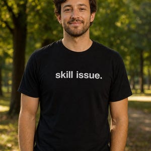Peut inclure: T-shirt noir à col rond avec le texte "skill issue." La chemise est portée par une personne debout à l'extérieur, les mains dans les poches. L'arrière-plan est une vue floue d'arbres.