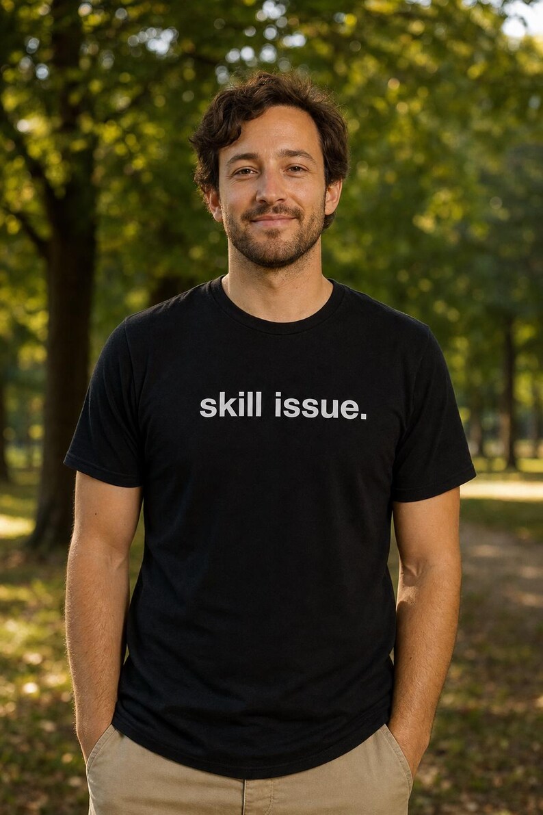 Peut inclure: T-shirt noir &agrave; col rond avec le texte "skill issue." La chemise est port&eacute;e par une personne debout &agrave; l'ext&eacute;rieur, les mains dans les poches. L'arri&egrave;re-plan est une vue floue d'arbres.