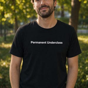 Op de afbeelding: Zwart T-shirt met ronde hals en de tekst "Permanent Underclass" in het wit. Het shirt wordt gedragen door een persoon die buiten staat met een vage groene achtergrond.