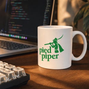 Peut inclure: Un mug en céramique blanche orné d'un motif graphique vert représentant un personnage jouant de la flûte, accompagné du texte "pied piper" en police serif verte. Le mug présente une forme cylindrique avec une anse incurvée.