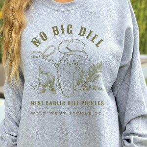Peut inclure: Un sweat-shirt gris clair avec un motif vert d'un cornichon souriant portant un chapeau de cow-boy, tenant un lasso, et le texte "NO BIG DILL". Les mots "MINI GARLIC DILL PICKLES" et "WILD WEST PICKLE CO." sont en dessous.