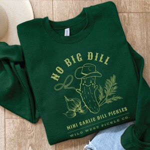 Peut inclure: Un sweat-shirt vert fonc&eacute; avec un motif vert clair. Le motif repr&eacute;sente un cornichon souriant portant un chapeau de cow-boy, un lasso et le texte "NO BIG DILL" et "MINI GARLIC DILL PICKLES WILD WEST PICKLE CO."