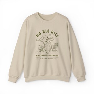 Peut inclure: Un sweat-shirt cr&egrave;me avec un dessin d'un cornichon souriant portant un chapeau de cowboy, un lasso et des gousses d'ail. Le texte dit "NO BIG DILL" et "MINI GARLIC DILL PICKLES" avec "WILD WEST PICKLE CO." en dessous.