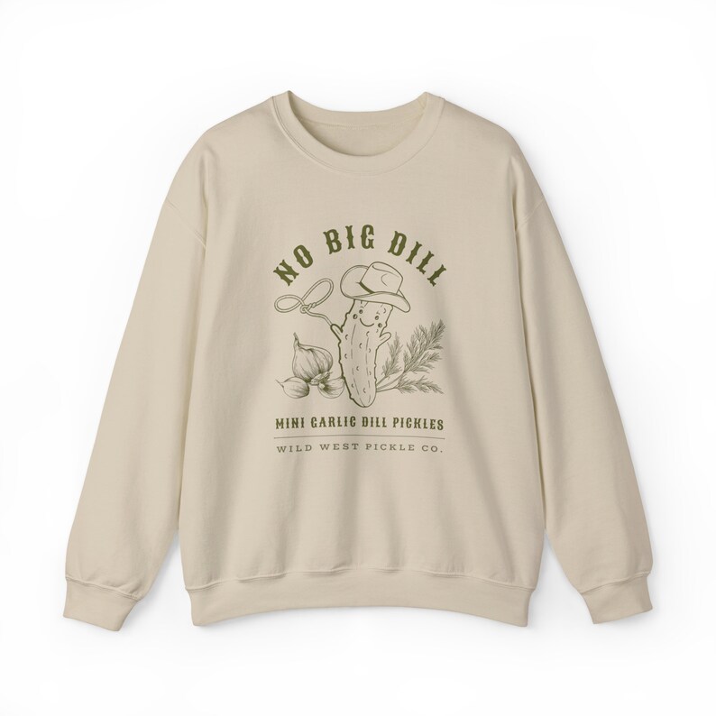 Peut inclure: Un sweat-shirt cr&egrave;me avec un dessin d'un cornichon souriant portant un chapeau de cowboy, un lasso et des gousses d'ail. Le texte dit "NO BIG DILL" et "MINI GARLIC DILL PICKLES" avec "WILD WEST PICKLE CO." en dessous.