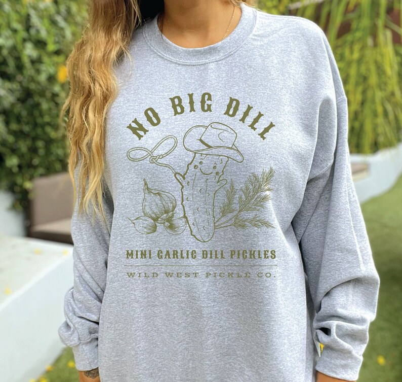 Peut inclure: Un sweat-shirt gris clair avec un motif vert d'un cornichon souriant portant un chapeau de cow-boy, tenant un lasso, et le texte "NO BIG DILL". Les mots "MINI GARLIC DILL PICKLES" et "WILD WEST PICKLE CO." sont en dessous.