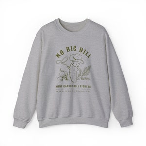 Peut inclure: Un sweat-shirt gris chin&eacute; avec un motif vert d'un cornichon souriant portant un chapeau de cowboy, un lasso et le texte "NO BIG DILL". Les mots "MINI GARLIC DILL PICKLES" et "WILD WEST PICKLE CO." sont en dessous.