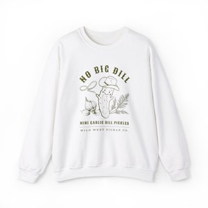 Peut inclure: Sweat-shirt blanc avec un dessin vert d'un cornichon souriant portant un chapeau de cow-boy, un lasso et de l'ail. Le texte indique "NO BIG DILL" et "MINI GARLIC DILL PICKLES WILD WEST PICKLE CO."