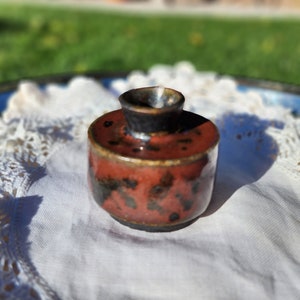 Könnte beinhalten: Eine kleine, runde, rot-braune Keramikvase mit schwarzen Flecken. Die Vase hat eine schmale Öffnung oben.