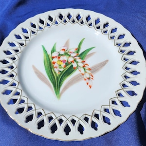 May include: White porcelain plate with a delicate openwork design around the edge. The center features a hand-painted floral design of white and orange flowers with green leaves.