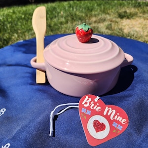 Puede incluir: Un horneador de brie rosa con un pomo en forma de fresa en la tapa, acompañado de una espátula de madera. Una etiqueta en forma de corazón con las palabras "Brie Mine" está adjunta. El conjunto está sobre una superficie azul.