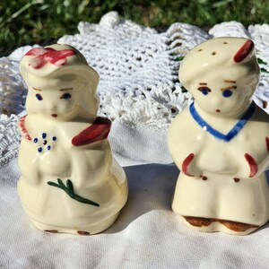Puede incluir: Un juego de salero y pimentero vintage con una niña y un niño con un atuendo de marinero. La niña lleva un vestido rojo y blanco con una flor azul y un lazo rojo. El niño lleva un atuendo de marinero azul y blanco con un sombrero rojo. Ambas figuras están pintadas de blanco con detalles azules y rojos.