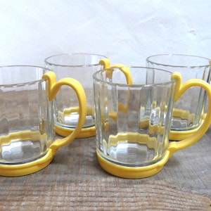 May include: Set of four clear glass mugs with yellow plastic handles and bases. The mugs have a ribbed design.
