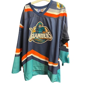 Könnte beinhalten: Marineblaues Hockey-Trikot mit türkisen, orangefarbenen und weißen Akzenten. Das Trikot hat ein großes Logo mit dem Wort "Islanders" und eine Figur in einem Kapuzenmantel, die einen Hockeyschläger hält.