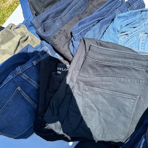 May include: A pile of various denim jeans in shades of blue, black, and camouflage. The jeans are folded and stacked, showcasing different styles and washes. Some jeans have visible back pockets and belt loops.