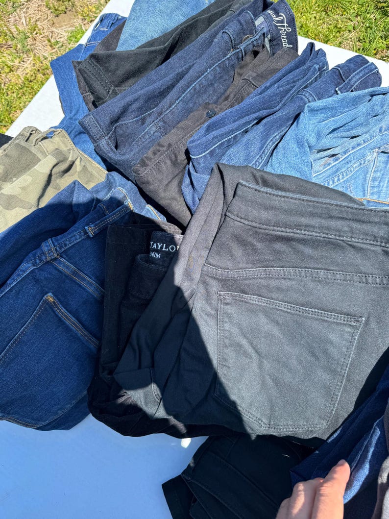 May include: A pile of various denim jeans in shades of blue, black, and camouflage. The jeans are folded and stacked, showcasing different styles and washes. Some jeans have visible back pockets and belt loops.