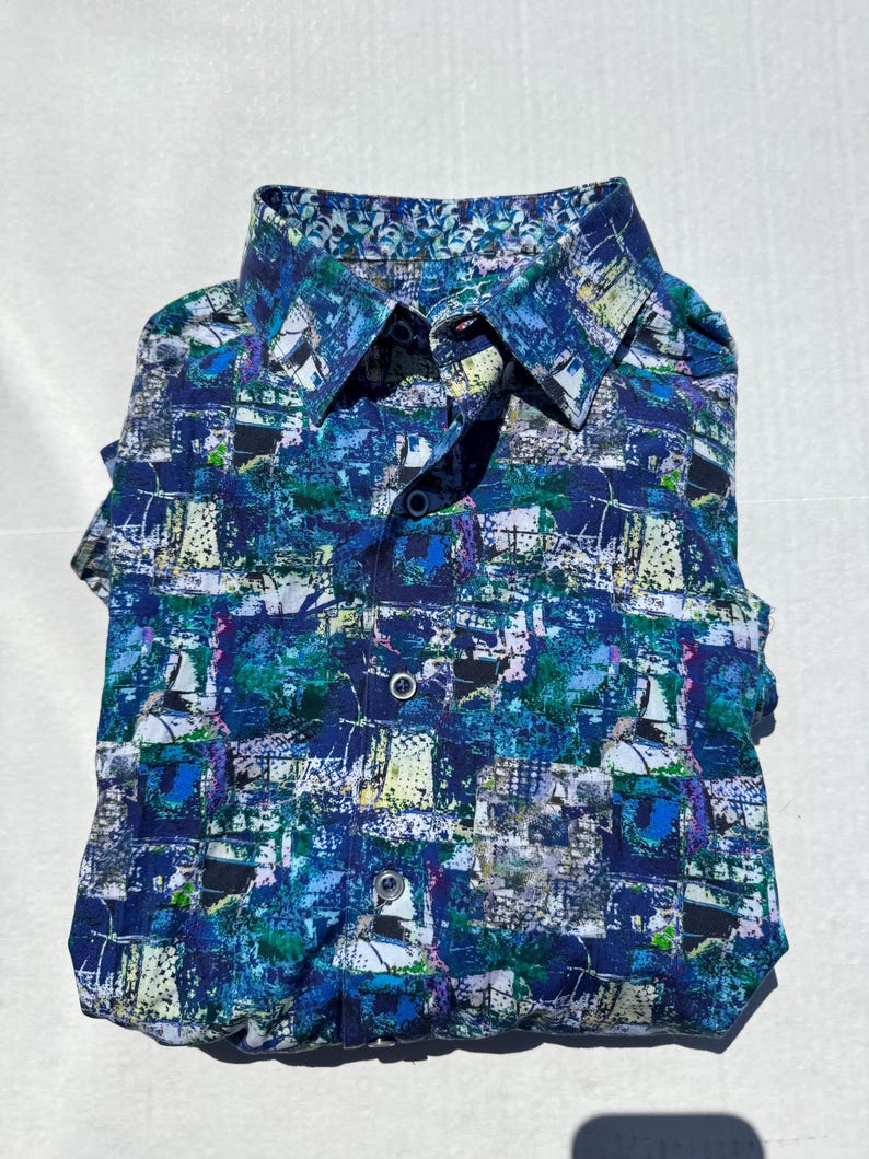 May include: Folded button-down shirt with a vibrant abstract pattern in shades of blue, green, and white. The shirt features a contrasting patterned collar and cuffs. The design includes a button-down front and short sleeves.