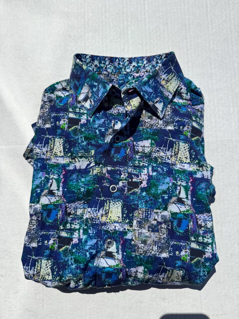 May include: A folded button-down shirt with a vibrant, abstract pattern in shades of blue, green, and white. The design includes what appears to be sailboat imagery. The shirt has a classic collar and button closure.