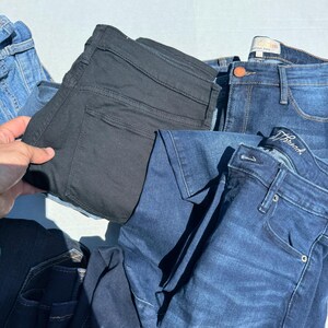May include: A collection of denim jeans in various shades of blue and black. The jeans are folded and stacked, showcasing different styles and washes. The image is taken outdoors, with a glimpse of green grass in the background.